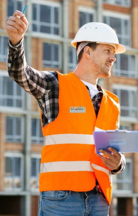 Ingeniero de construcción con chaleco de seguridad naranja y casco blanco de CUBIERFAC, sosteniendo documentos y señalando mientras inspecciona un edificio en construcción.