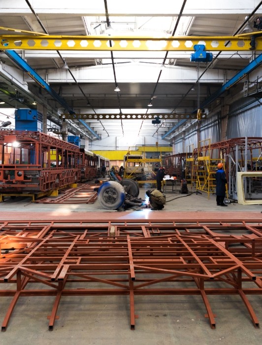 Interior de una planta industrial de CUBIERFAC SAS donde varios trabajadores realizan labores de soldadura y ensamblaje de estructuras metálicas pintadas de rojo, con grúas y maquinaria pesada en el techo y laterales del taller.